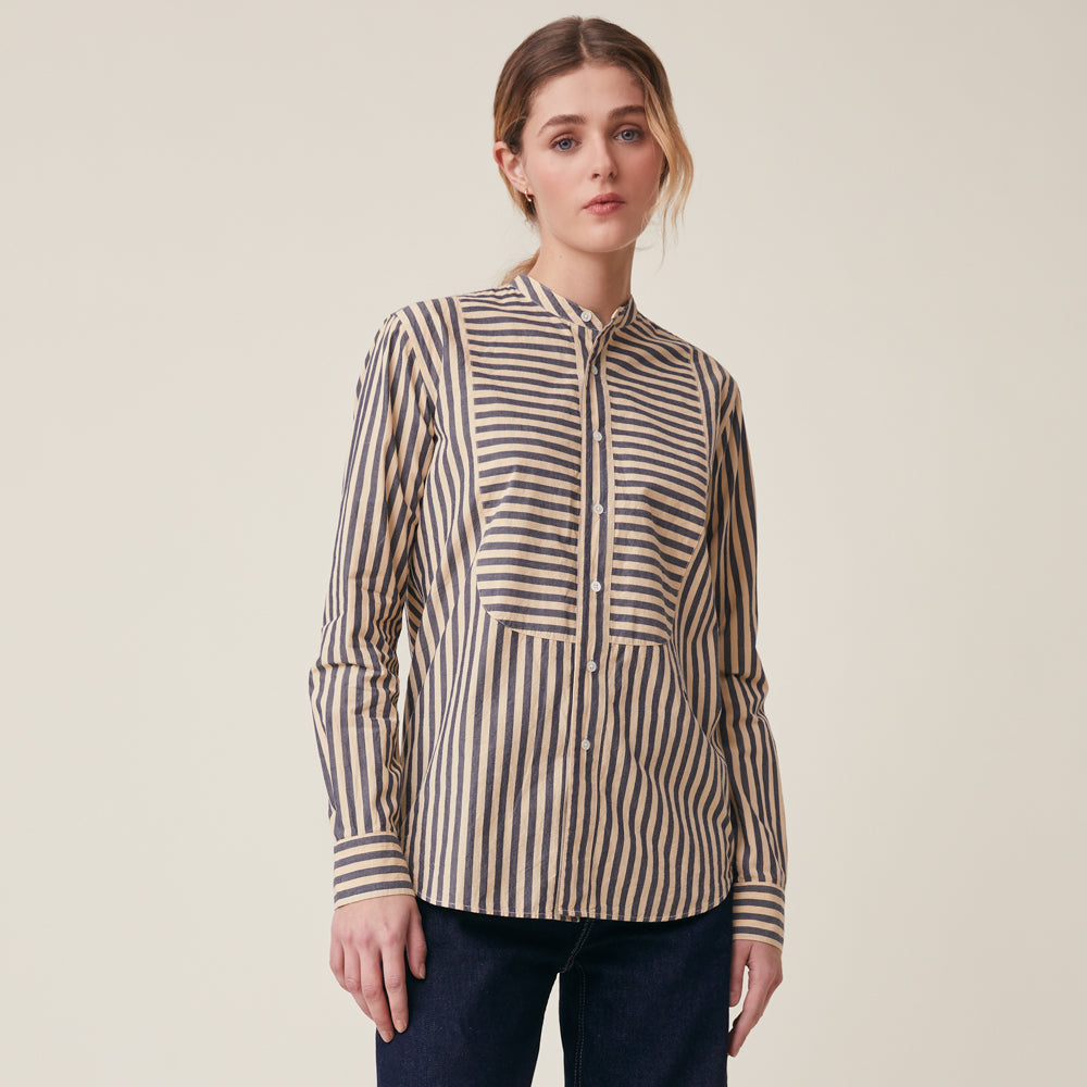 A woman modeling a beige and navy vertically striped long-sleeve button-up shirt with a mandarin collar, photographed against a solid light beige studio background.