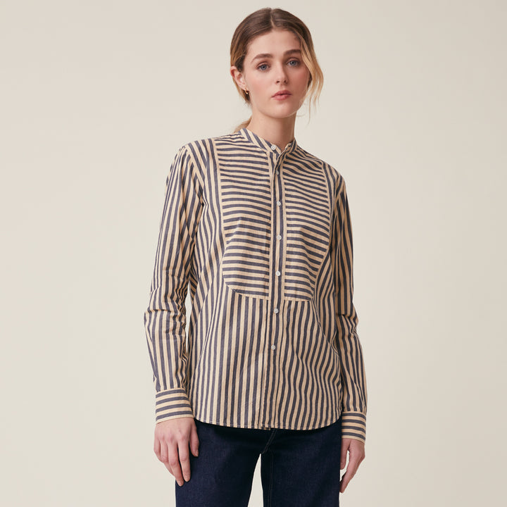 A woman modeling a beige and navy vertically striped long-sleeve button-up shirt with a mandarin collar, photographed against a solid light beige studio background.
