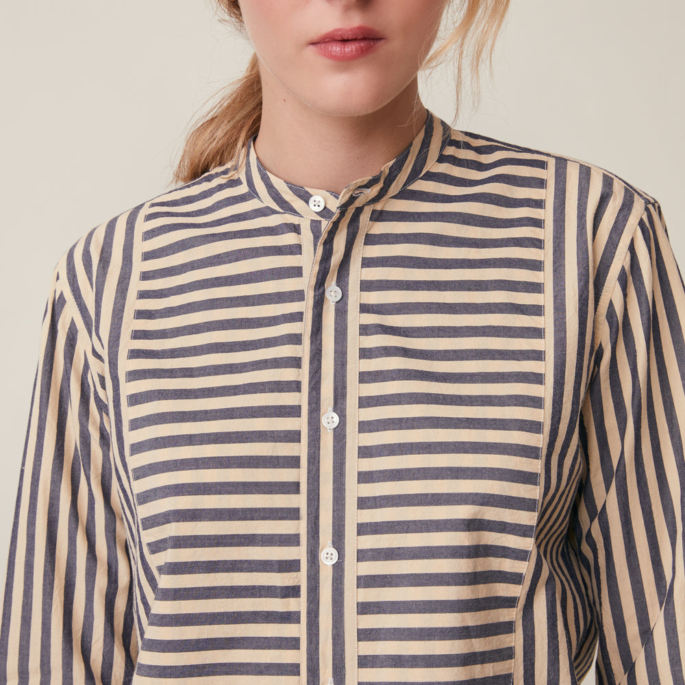 A close-up of a woman wearing a long-sleeve button-front shirt with narrow horizontal stripes in navy and warm beige, against a plain light beige background.
