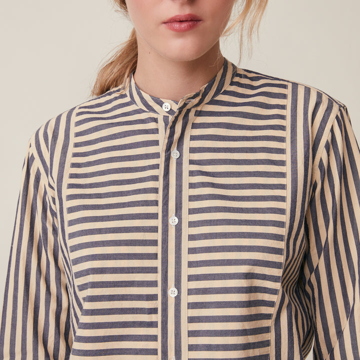 A close-up of a woman wearing a long-sleeve button-front shirt with narrow horizontal stripes in navy and warm beige, against a plain light beige background.