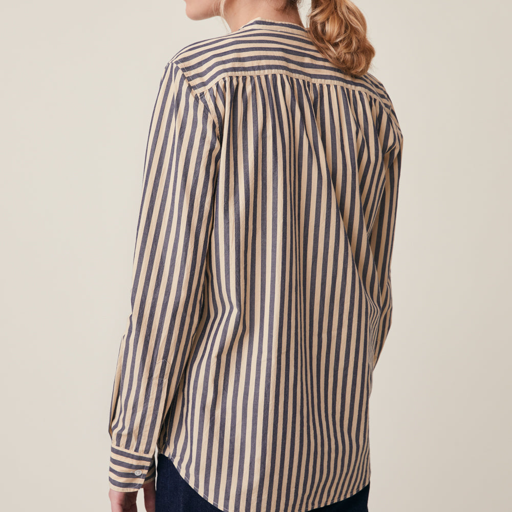 A woman wearing a long-sleeve button-up shirt with vertical navy and beige stripes, shown from the back against a plain light beige background.