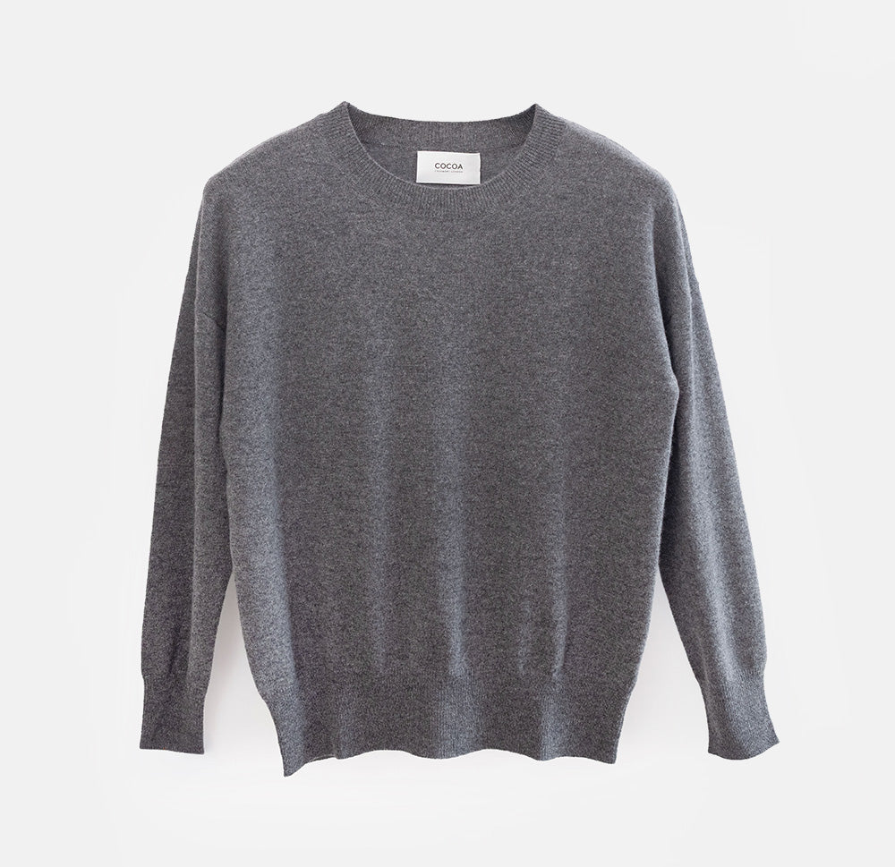 A dark gray long-sleeve knit sweater with a crew neck, laid flat against a light gray background. A small white brand label is visible at the inner collar.
