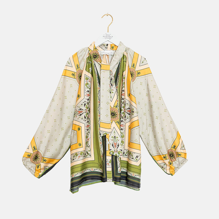 A long-sleeve pussy bow blouse with a cream base, yellow geometric borders, and green floral patterns, hanging on a white hanger against a light gray background.