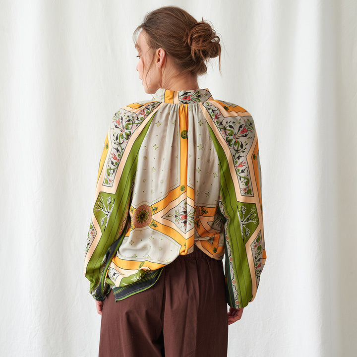 A woman shown from the back wearing a long-sleeve silk blouse with a vibrant retro geometric and floral print in green, yellow, orange, and beige, paired with brown trousers against a white curtain background.