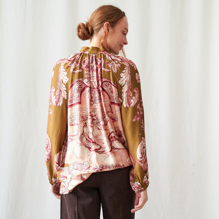 A woman shown from the back wearing a long-sleeve silk blouse with a gold-ochre base and a pink and beige baroque-style floral pattern, paired with dark brown trousers, set against a plain white wall.