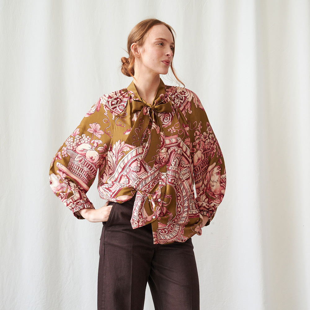 A woman modeling a long-sleeve blouse with a brown base and a pink and red paisley pattern, paired with dark brown trousers, standing against a white curtain backdrop.