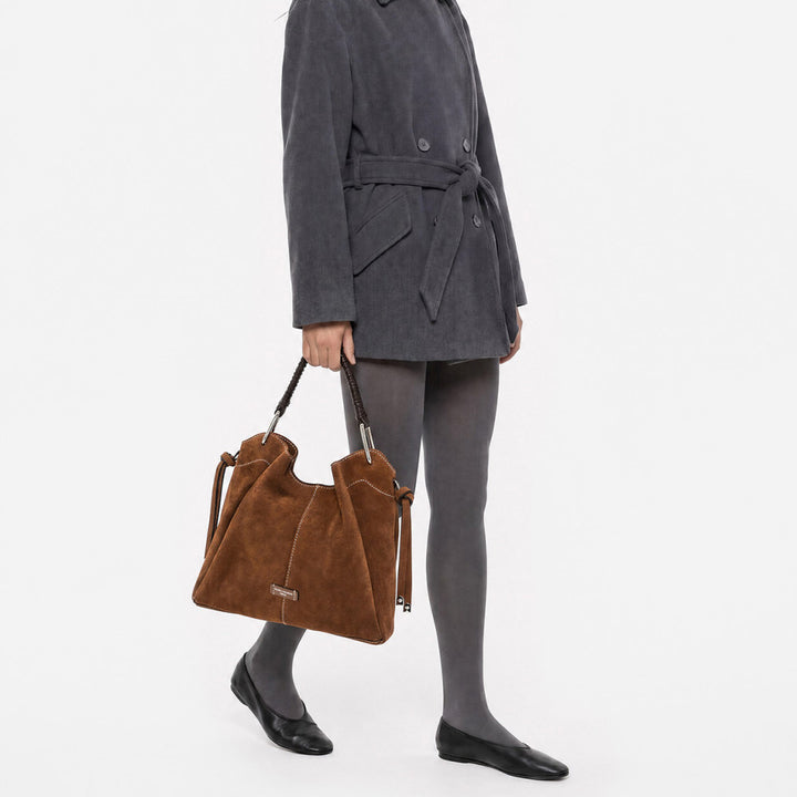 Woman in a grey coat carrying a large tan suede bag by its braided handle
