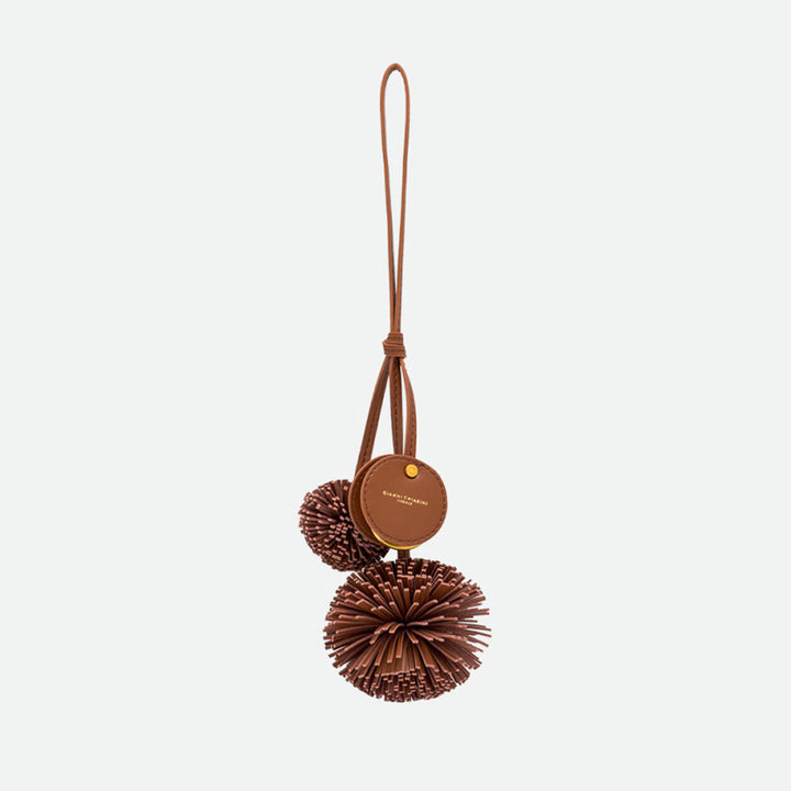 Brown bag charm with two fringed pompoms, a round branded disc and a long loop