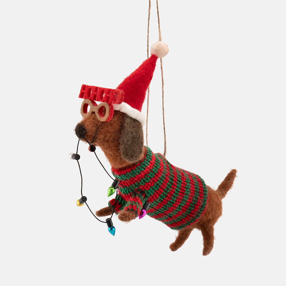 A felt dachshund Christmas tree decoration wearing a red and green striped sweater, a red Santa hat, and novelty glasses, with a string of holiday lights around its neck, shown against a light gray background.