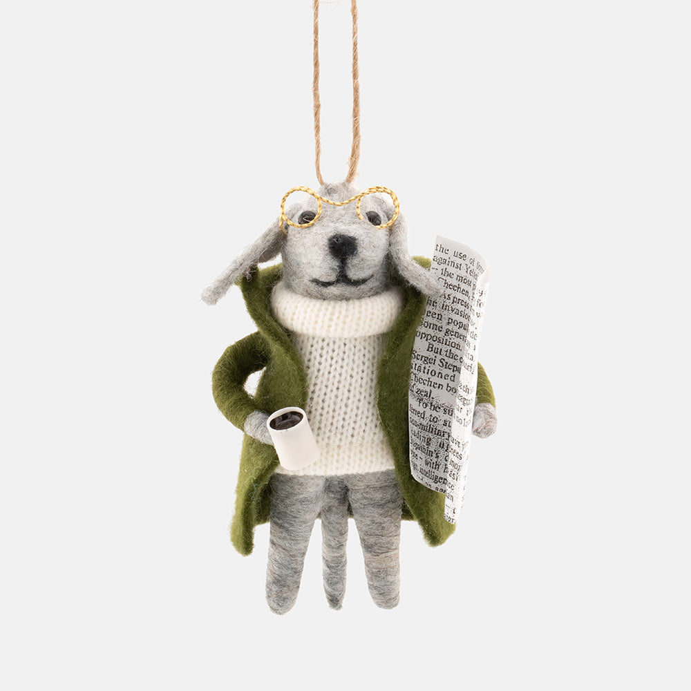 A felted wool dog Christmas tree decoration dressed in a green coat, cream knit sweater, and gold-rimmed glasses, holding a rolled-up newspaper, shown against a light gray background.