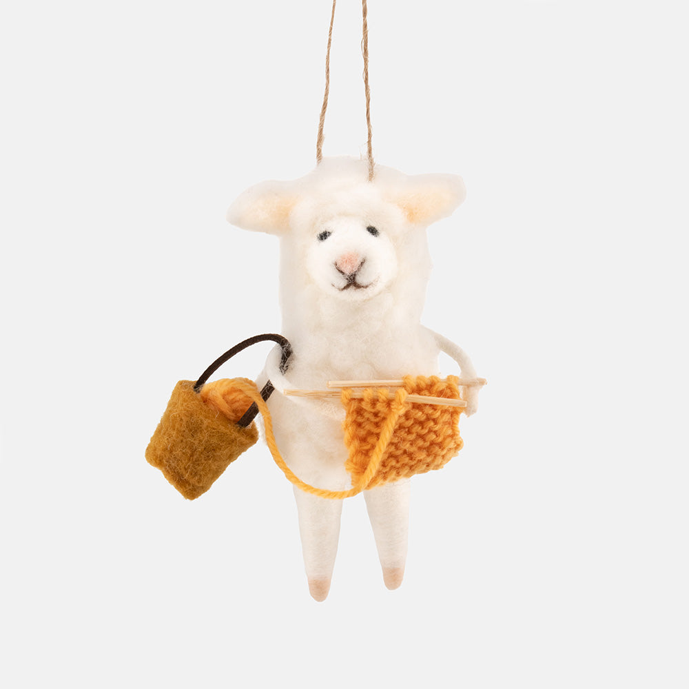 A white  felt sheep hanging christmas tree decoration holding a small orange knitted pouch and knitting needles, suspended by a string against a light gray background.