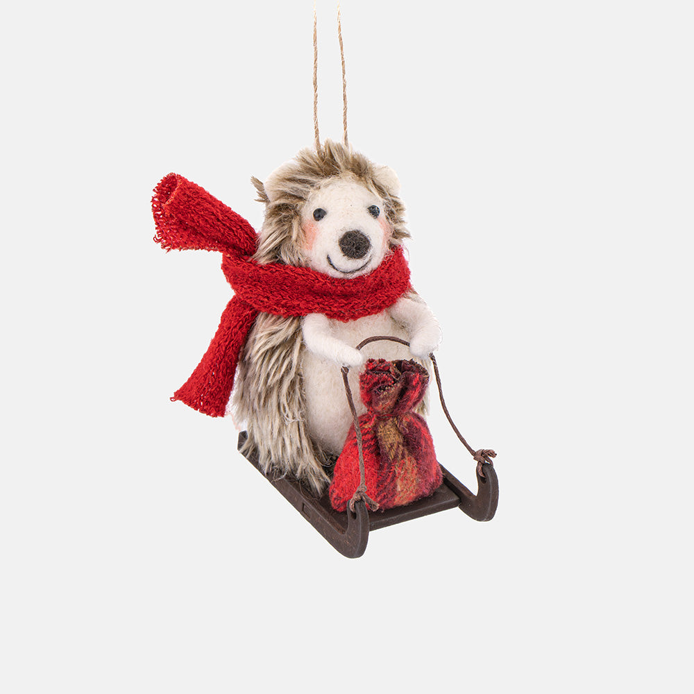 A plush hedgehog Christmas tree decoration sitting in a small sled, wearing a bright red knit scarf and holding a red patterned sack, photographed against a plain light gray background.