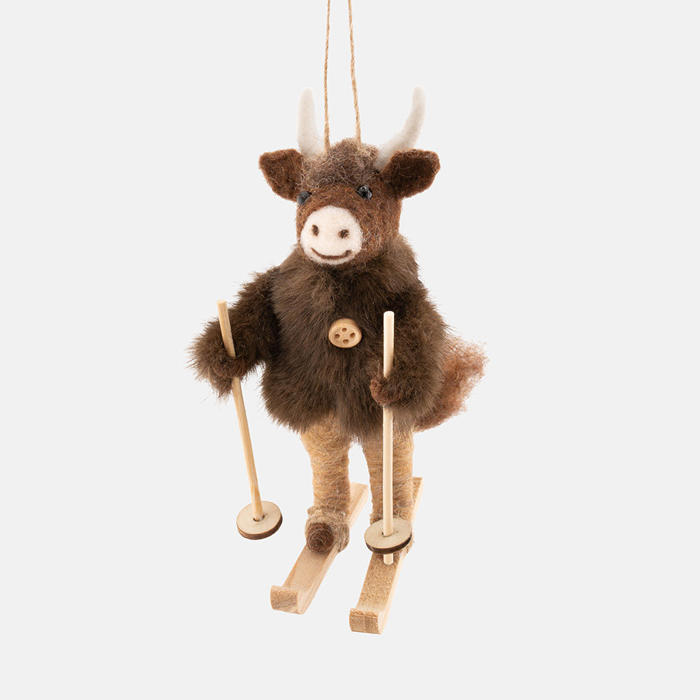A plush Highland cow hanging christmas tree decoration with brown fur, with white horns, a wooden button chest detail, and wooden skis with poles, shown against a light gray background.
