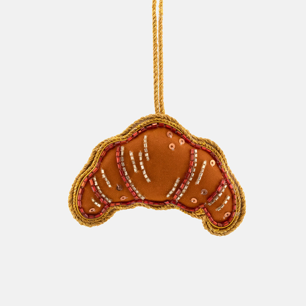 A croissant-shaped fabric christmas tree decoration in warm brown and orange tones, outlined with gold braided trim and decorated with small metallic bead details, suspended against a plain light gray background.
