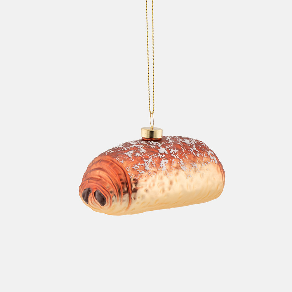 A pain au chcolat shaped hanging ornament with a glossy finish, dusted with white powdered sugar, suspended by a gold-coloured string against a light grey background.