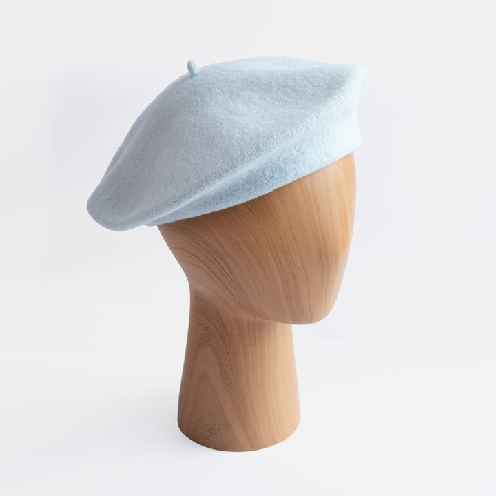 A light blue pink felt beret hat displayed on a light wood mannequin head against a clean white background.