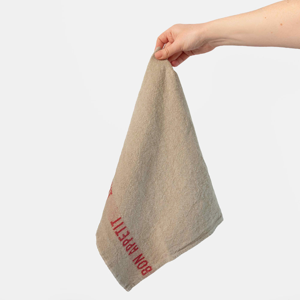 Hand holding a beige towel with red 'bon appetit' text on a white background