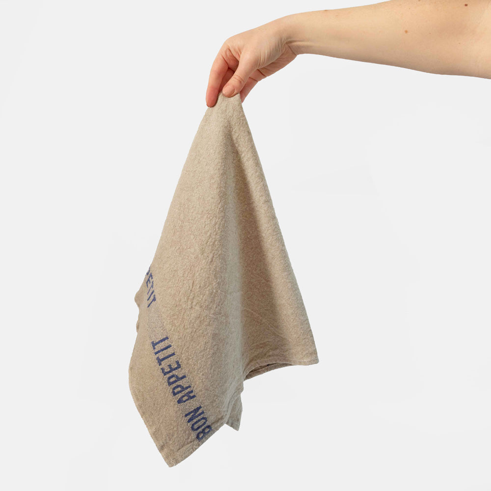 Beige tea towel with blue 'Bon Appetit' text held by a hand on a light grey background