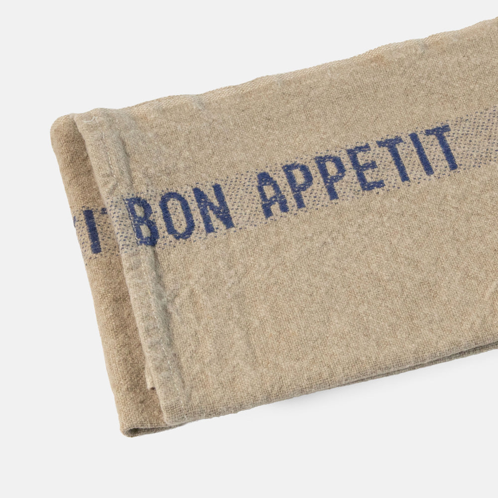 Beige tea towel with blue 'Bon Appetit' text held by a hand on a light grey background