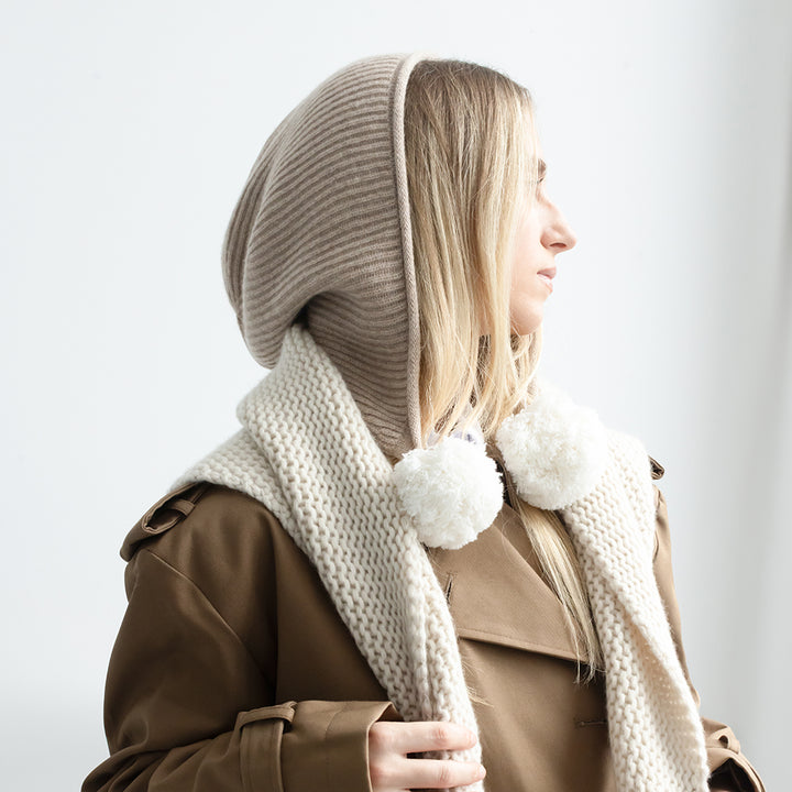 A woman in profile wearing a ribbed taupe knit bonnet with fluffy white pom-poms, layered over a chunky cream knit scarf and a brown trench coat against a light gray background.
