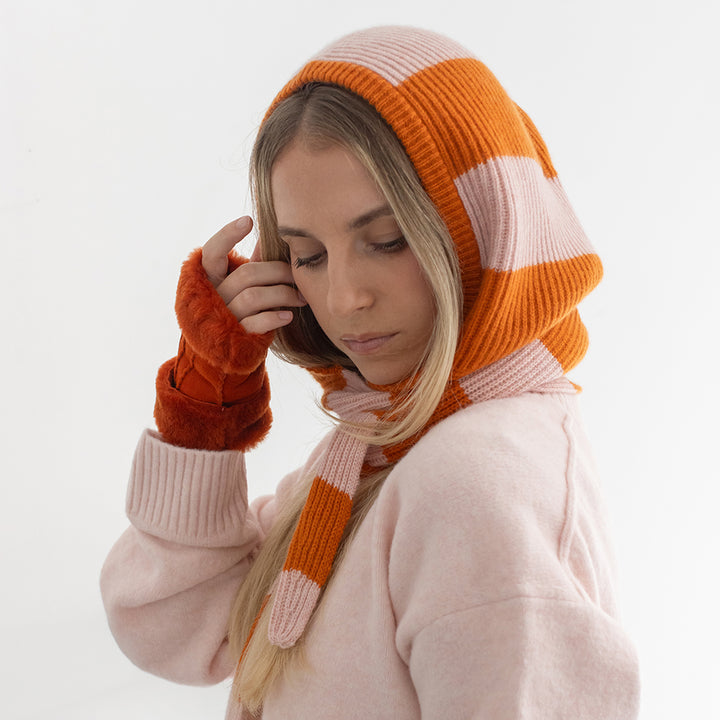 A woman wearing an orange and pink stripe knitted hooded scarf with orange leather mittens and light pink sweater against a plain white background.