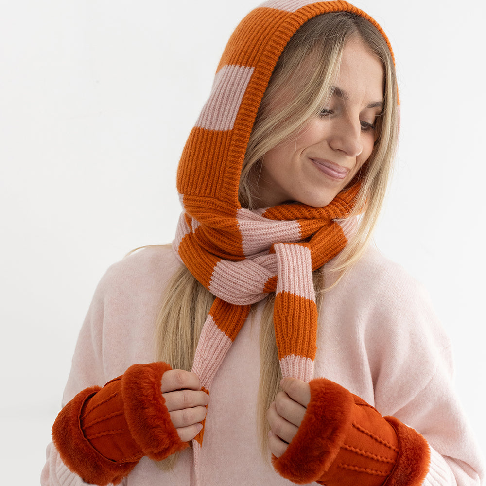 A woman wearing an orange and pink stripe knitted hooded scarf with orange leather mittens and light pink sweater against a plain white background.