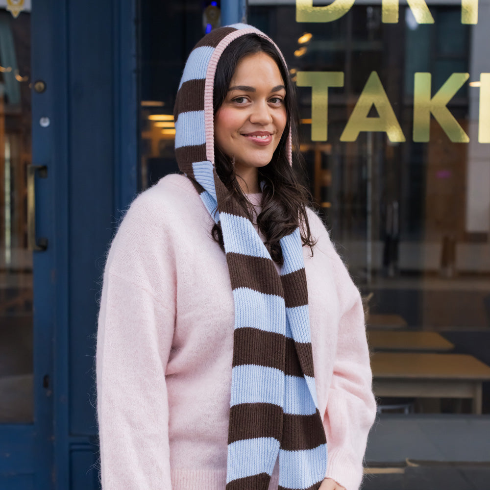 woman wearing a light pink wool sweater and a dark brown and light blue striped hooded scarf standing in front of a shop window