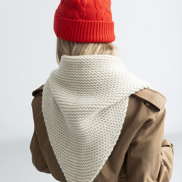 A person shown from the back wearing a chunky cream knit hooded cowl/scarf, a red knit beanie, and a brown trench coat against a plain light gray studio background.