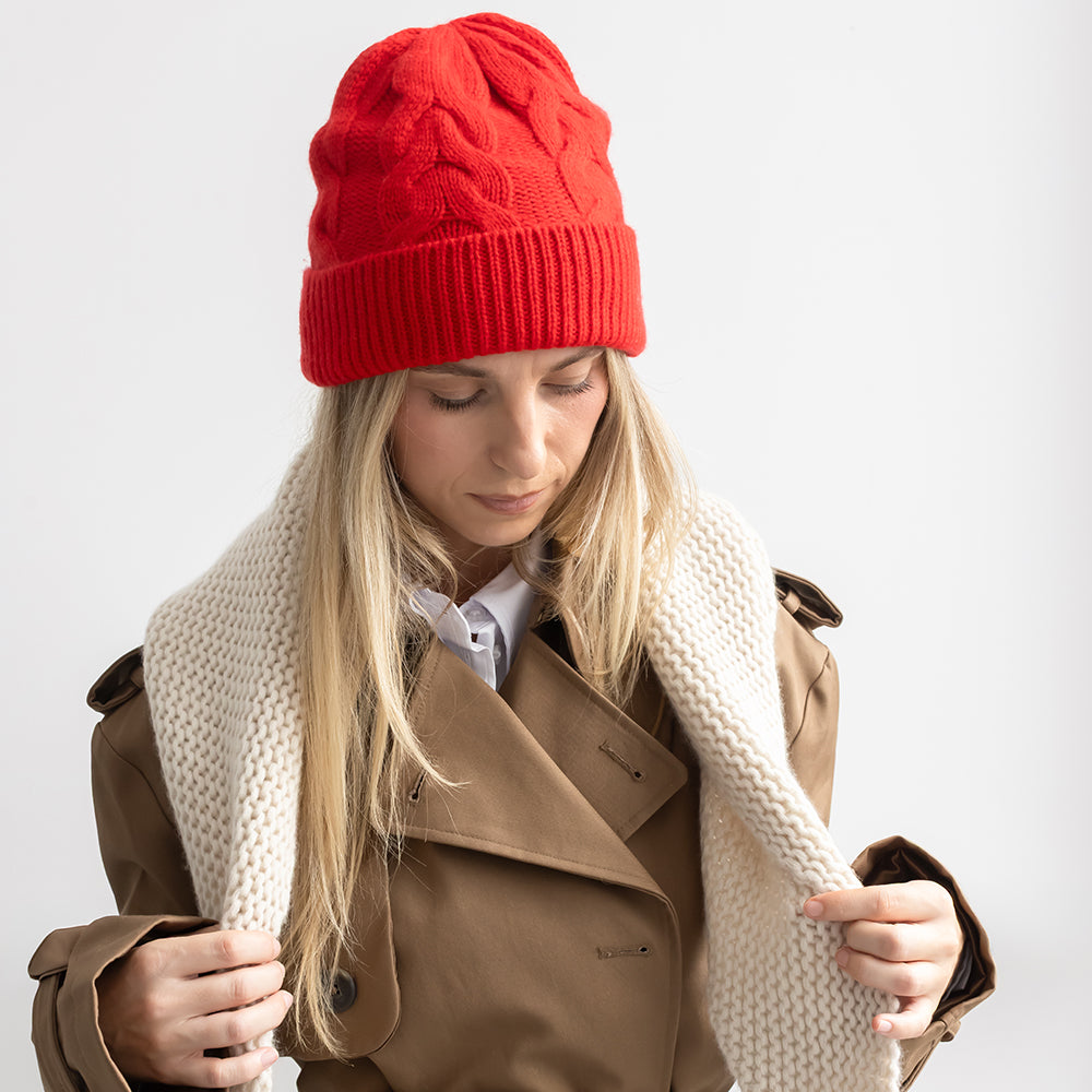 A woman wearing a bright red cable-knit beanie, a brown trench coat, and a chunky cream knit scarf, looking down against a light gray background.