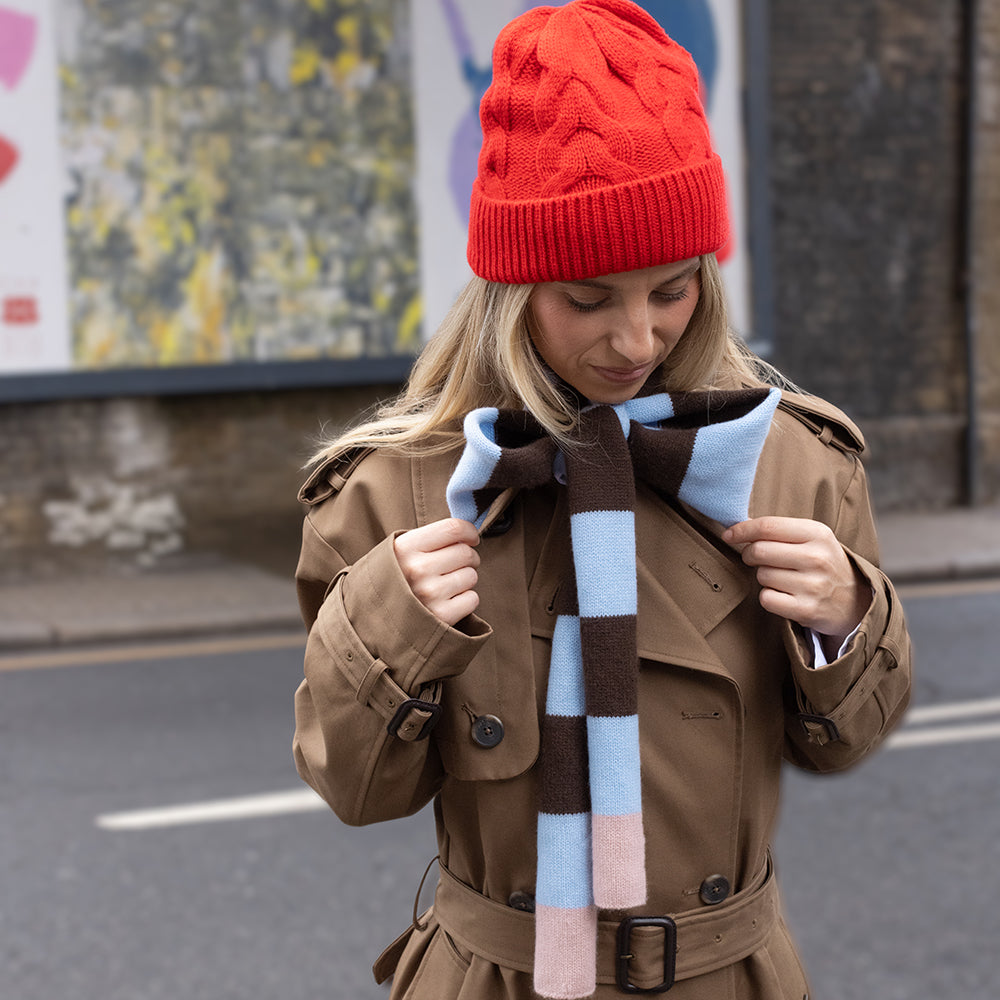 woman wearing a brown trench coat, red cable knit beanie hat and a brown and light blue stripe skinny scarf tied in a bow, in a street setting