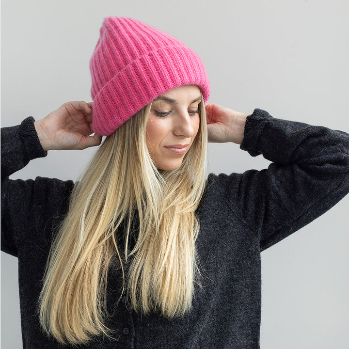 A woman with long blonde hair wearing a bright pink ribbed knit beanie and a dark gray cardigan, adjusting the beanie with both hands against a plain light gray background.