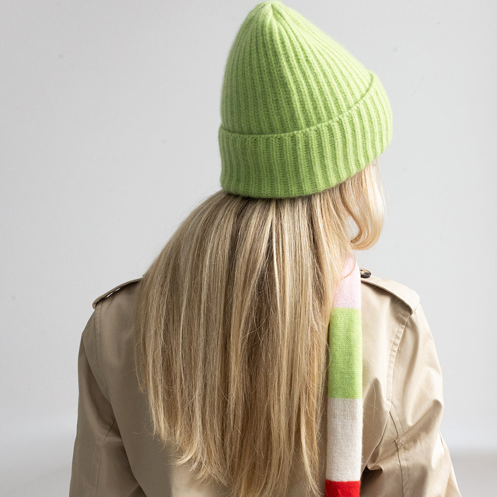 A person shown from the back wearing a bright lime green ribbed knit beanie, a beige trench coat, and a striped scarf in green, pink, white, and red, against a plain light gray background.