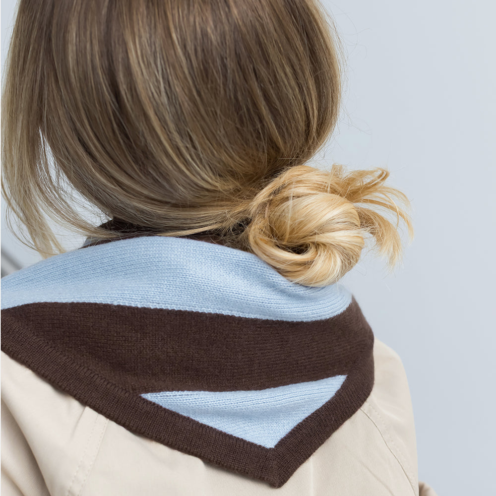 A close-up back view of a person wearing a beige coat with a knitted triangle shaped scarf in light blue and dark brown, styled with blonde hair in a low bun.