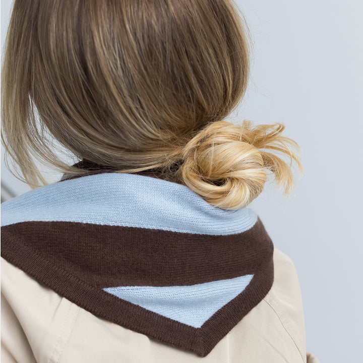 A close-up back view of a person wearing a beige coat with a knitted triangle shaped scarf in light blue and dark brown, styled with blonde hair in a low bun.