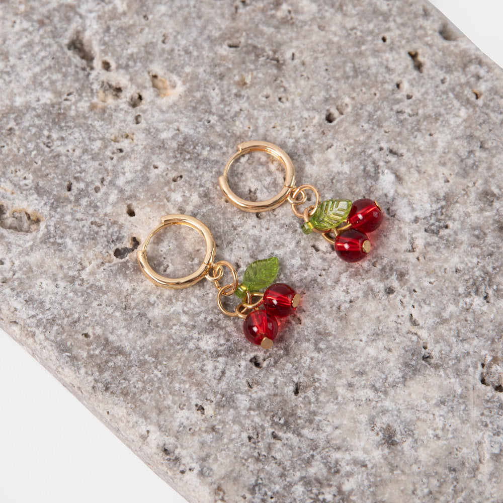 gold hoop earrings with red cherry shaped glass beads and green leaves on a textured stone background