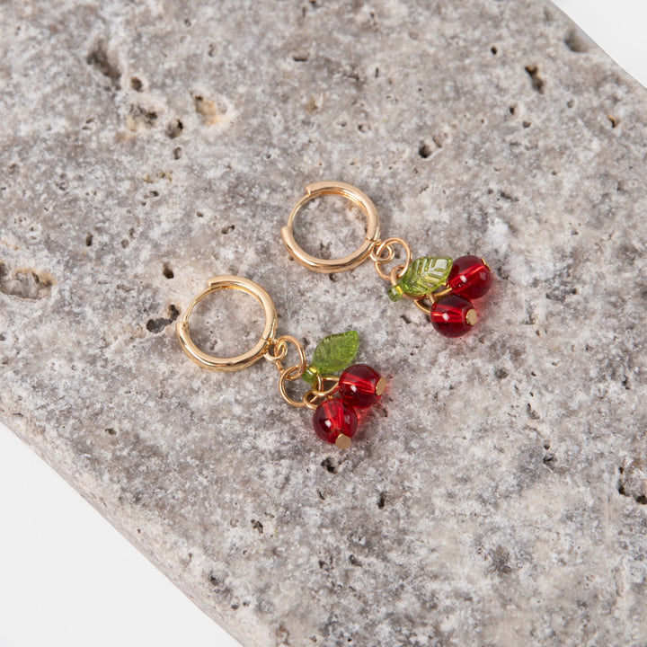 gold hoop earrings with red cherry shaped glass beads and green leaves on a textured stone background