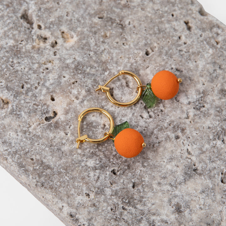 gold hoop earrings with orange shaped charms with green leaves on a textured stone background