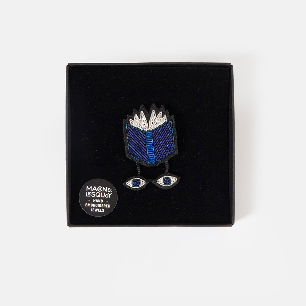 Hand embroidered brooch of an open blue book with silver pages and a pair of dangling embroidered eyes, on a white background.