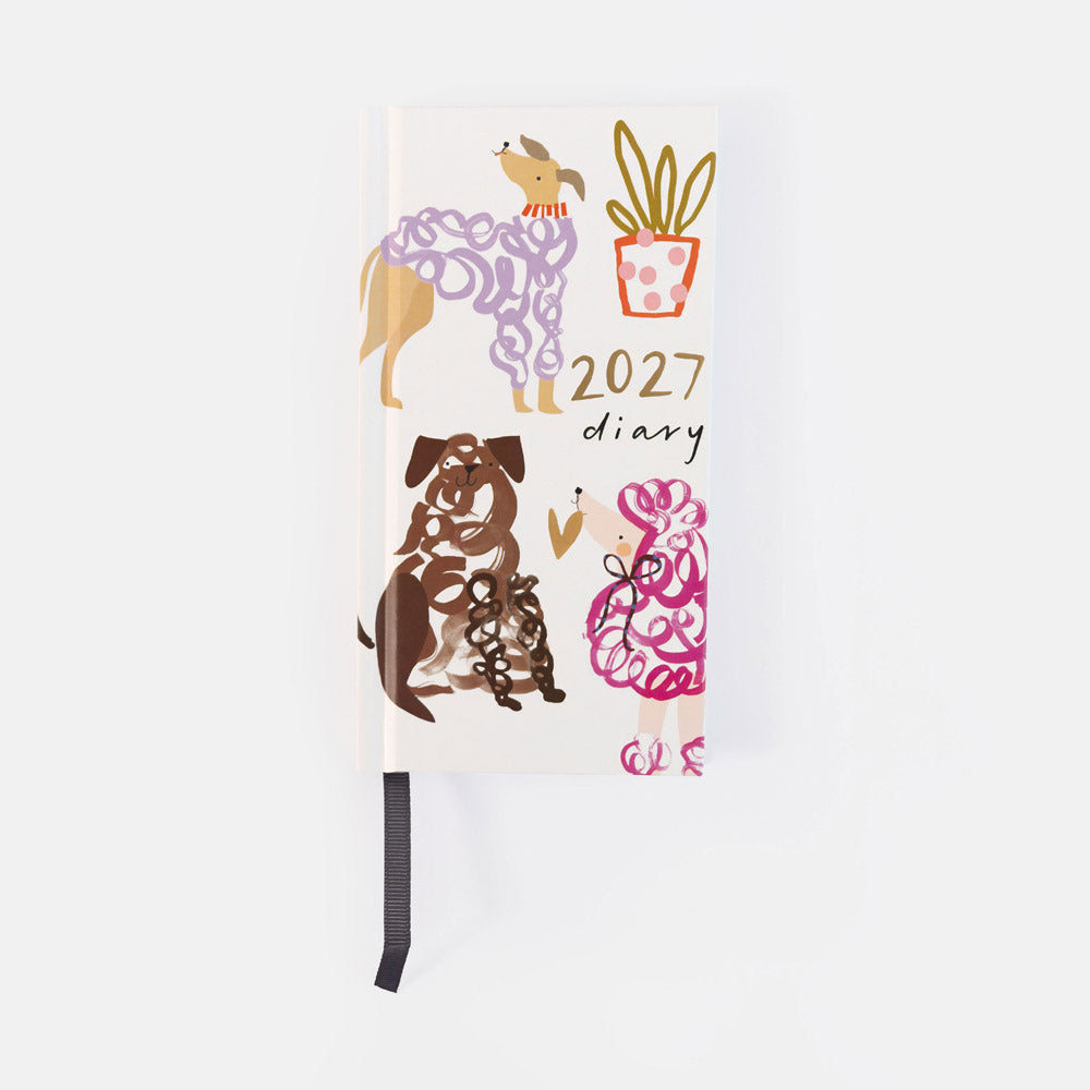 A 2027 daily planner with a white cover decorated with whimsical dog illustrations in purple, brown, and pink, along with a potted plant and gold text. A black ribbon bookmark hangs from the bottom.