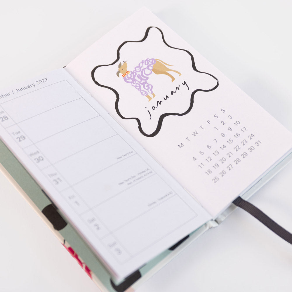 An open planner or notebook showing a stylized January calendar page with a black outline cloud, a purple patterned dress, and a small tan figure, photographed on a clean white background.