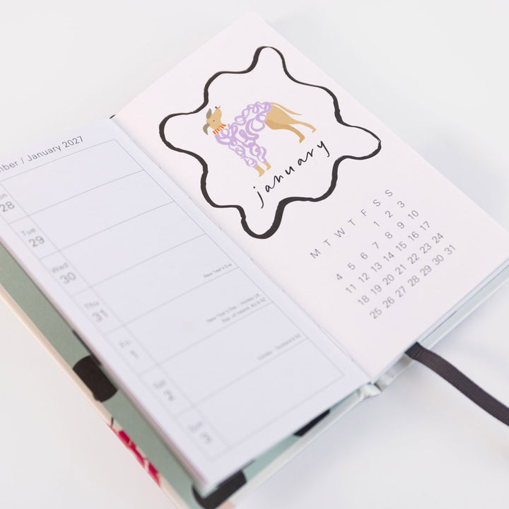 An open planner or notebook showing a stylized January calendar page with a black outline cloud, a purple patterned dress, and a small tan figure, photographed on a clean white background.