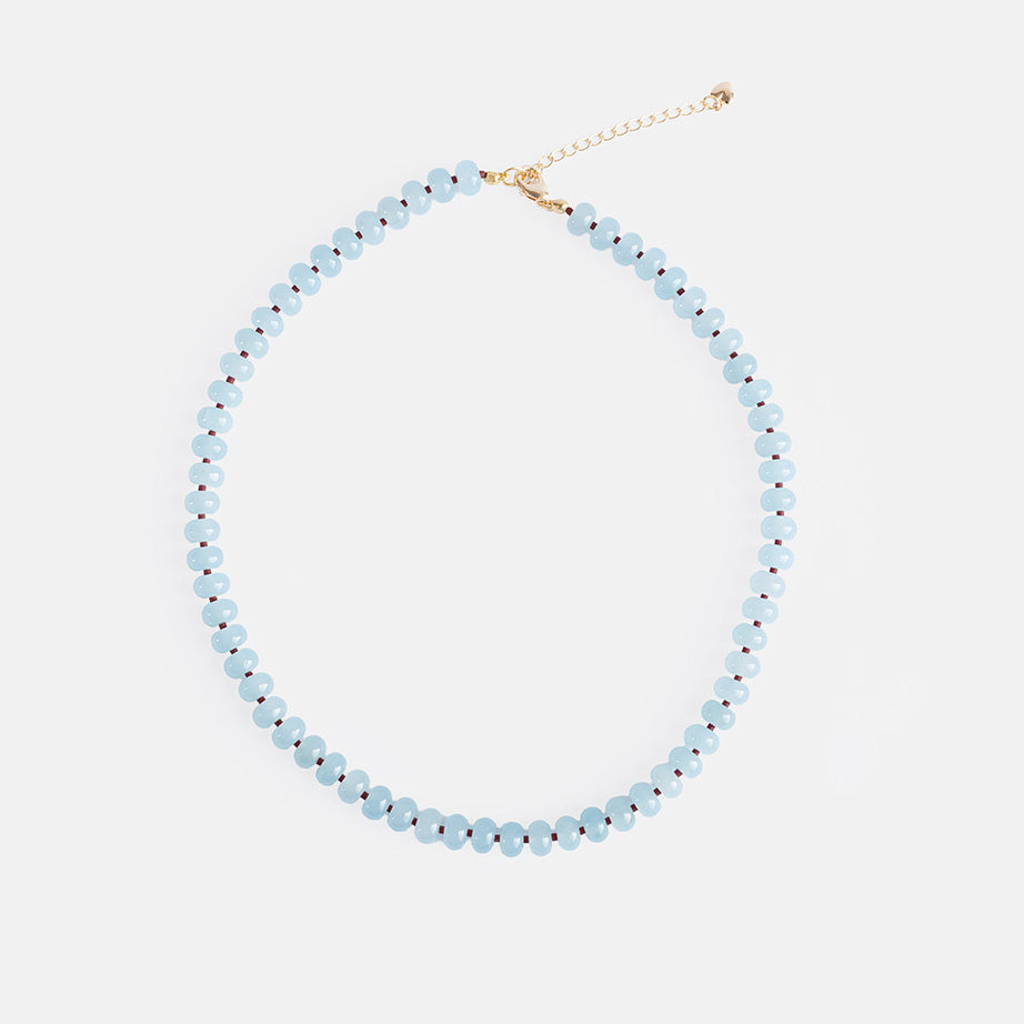 A handmade beaded necklace of pale blue semi-precious stone beads with tiny brown spacer beads and a gold-tone extender chain, on a light grey background.