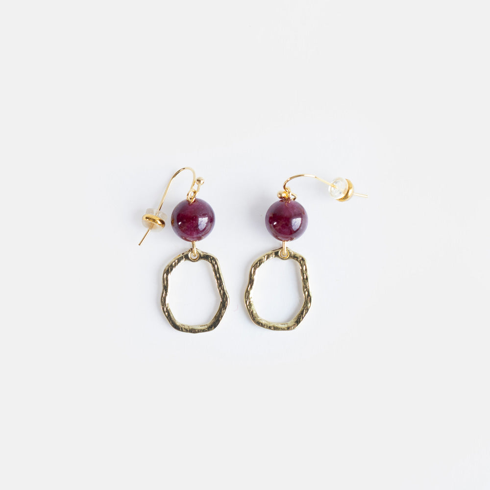 A pair of handmade drop earrings with a round burgundy semi-precious stone bead above an irregular antique gold-tone loop, on a light grey background.