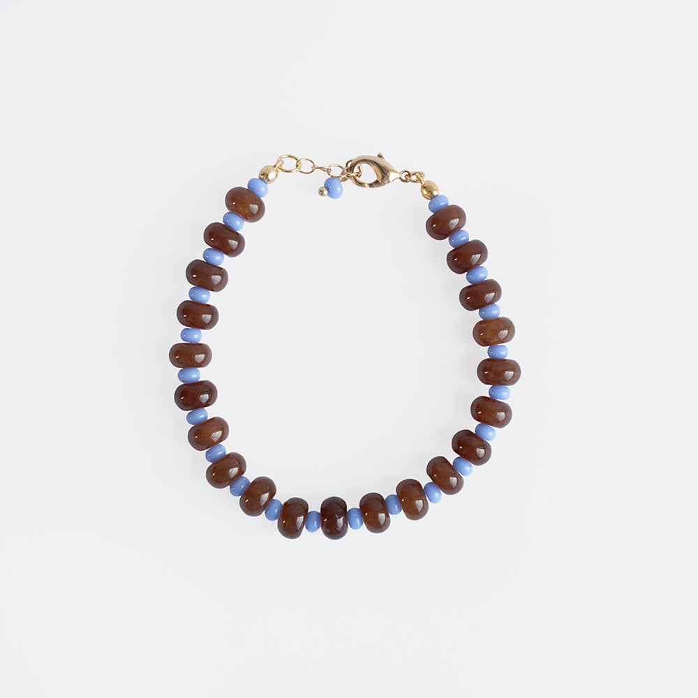 A handmade beaded bracelet of brown semi-precious stone rondelle beads alternating with periwinkle blue beads, with a gold-tone clasp and extender chain, on a light grey background.