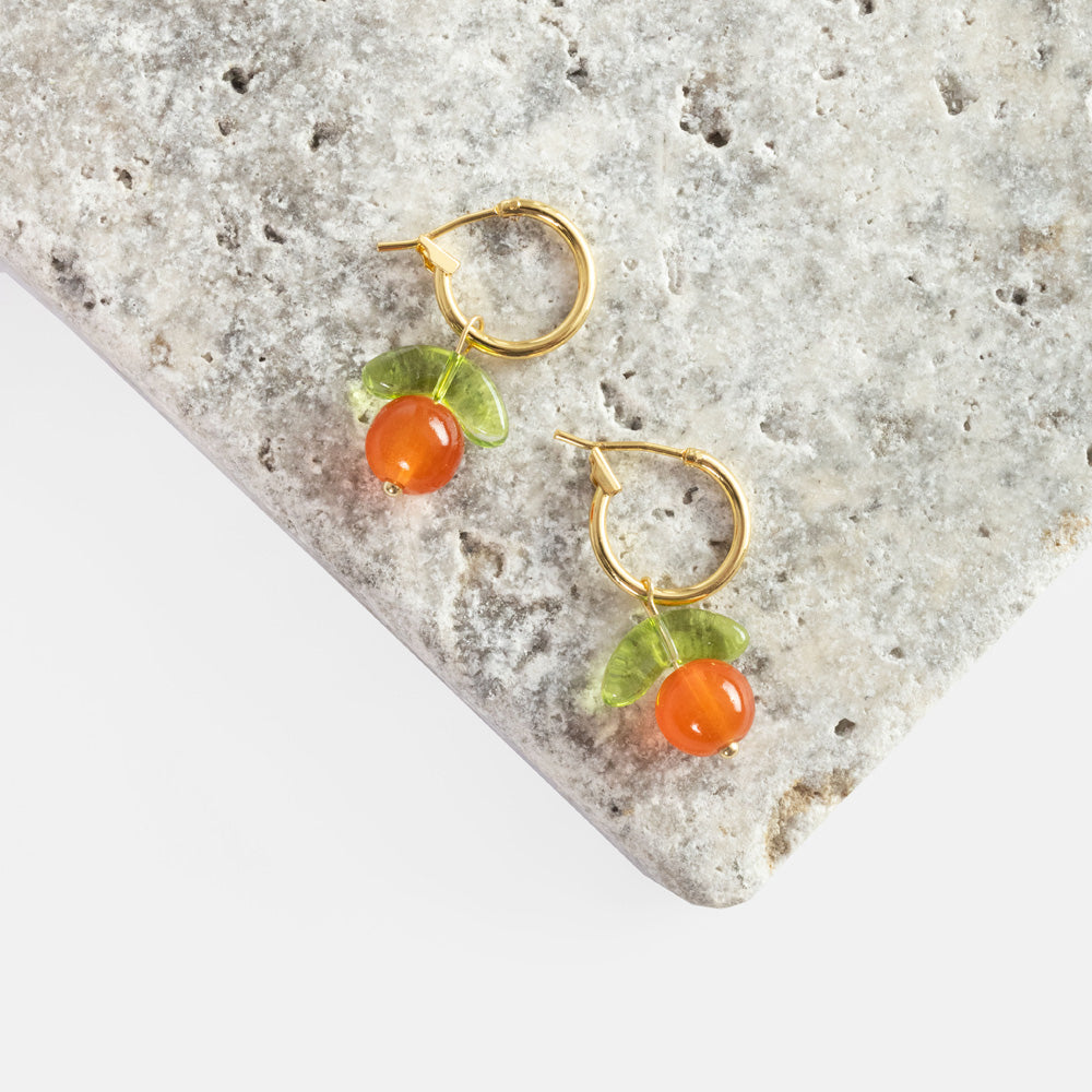 Gold hoop earrings with orange clementine glass bead drops and green leaves, styled on a textured stone surface.