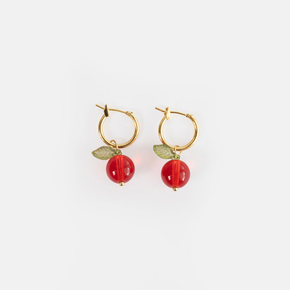 A pair of gold hoop earrings each with a glossy cranberry red glass bead and a green glass leaf, on a light grey background.