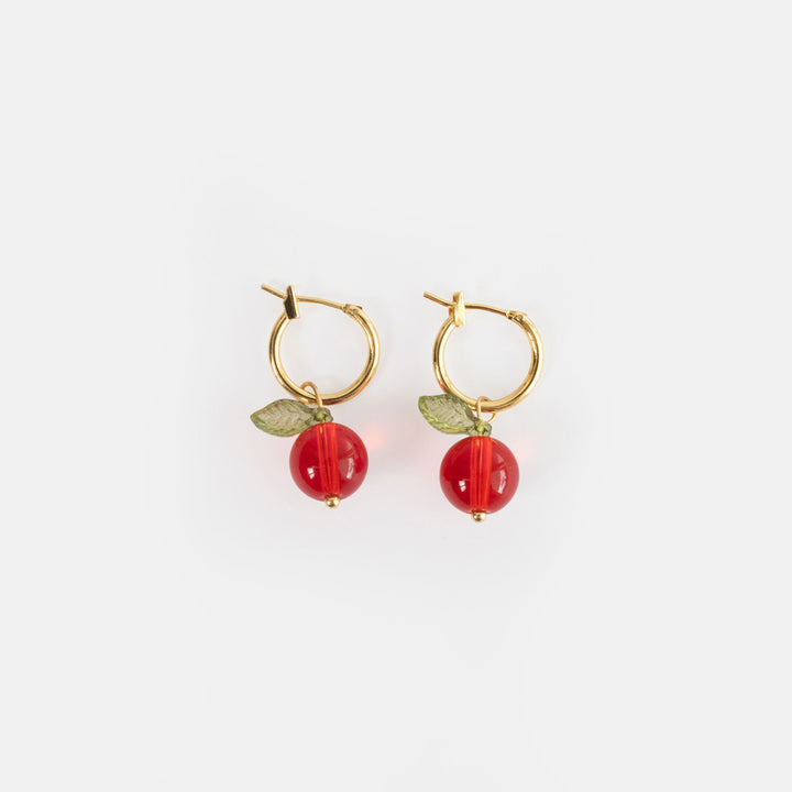 A pair of gold hoop earrings each with a glossy cranberry red glass bead and a green glass leaf, on a light grey background.