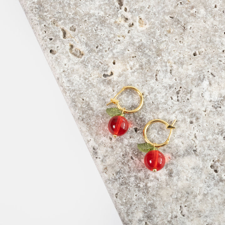 Gold hoop earrings with red cranberry glass bead drops and green leaves, styled on a textured stone surface.