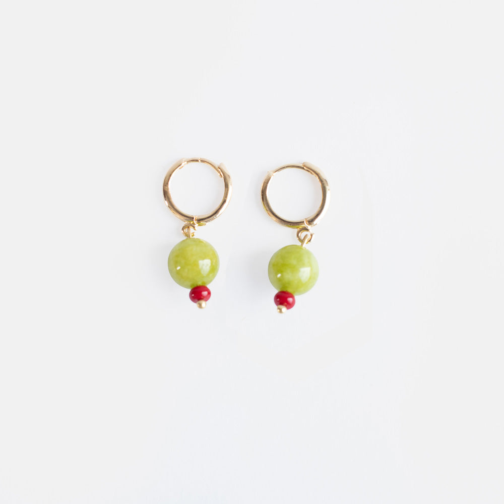 A pair of small gold huggie hoop earrings each with a round olive green semi-precious stone bead and a tiny red bead, on a light grey background.