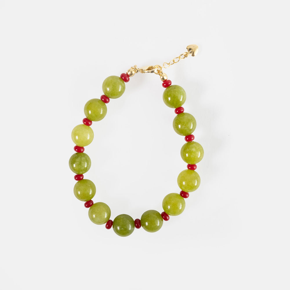 A handmade bracelet of round olive green semi-precious stone beads separated by small red beads, with a gold-tone clasp, extender chain and heart charm, on a light grey background.